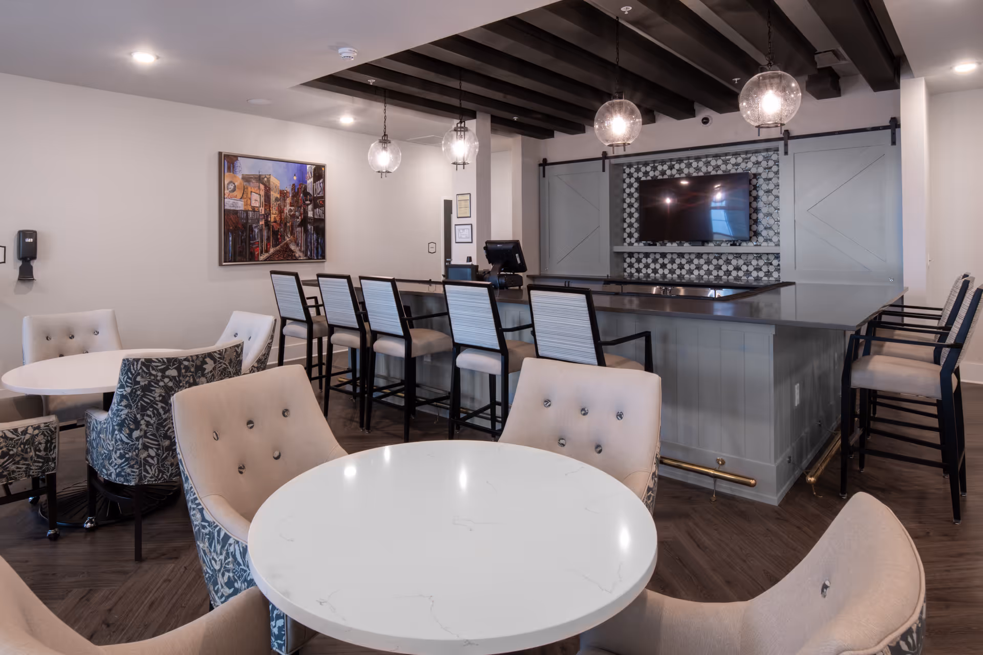 A modern common area with round white tables surrounded by cushioned chairs, a bar counter with high chairs, pendant lights hanging from a ceiling with exposed dark wooden beams, a wall-mounted TV, and a framed artwork on the wall.