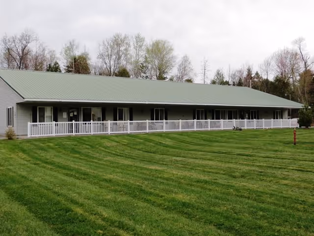 Single-story building with a green metal roof and light gray siding, featuring a long white railing along the front porch. The building is surrounded by a well-maintained green lawn and trees in the background under a cloudy sky.