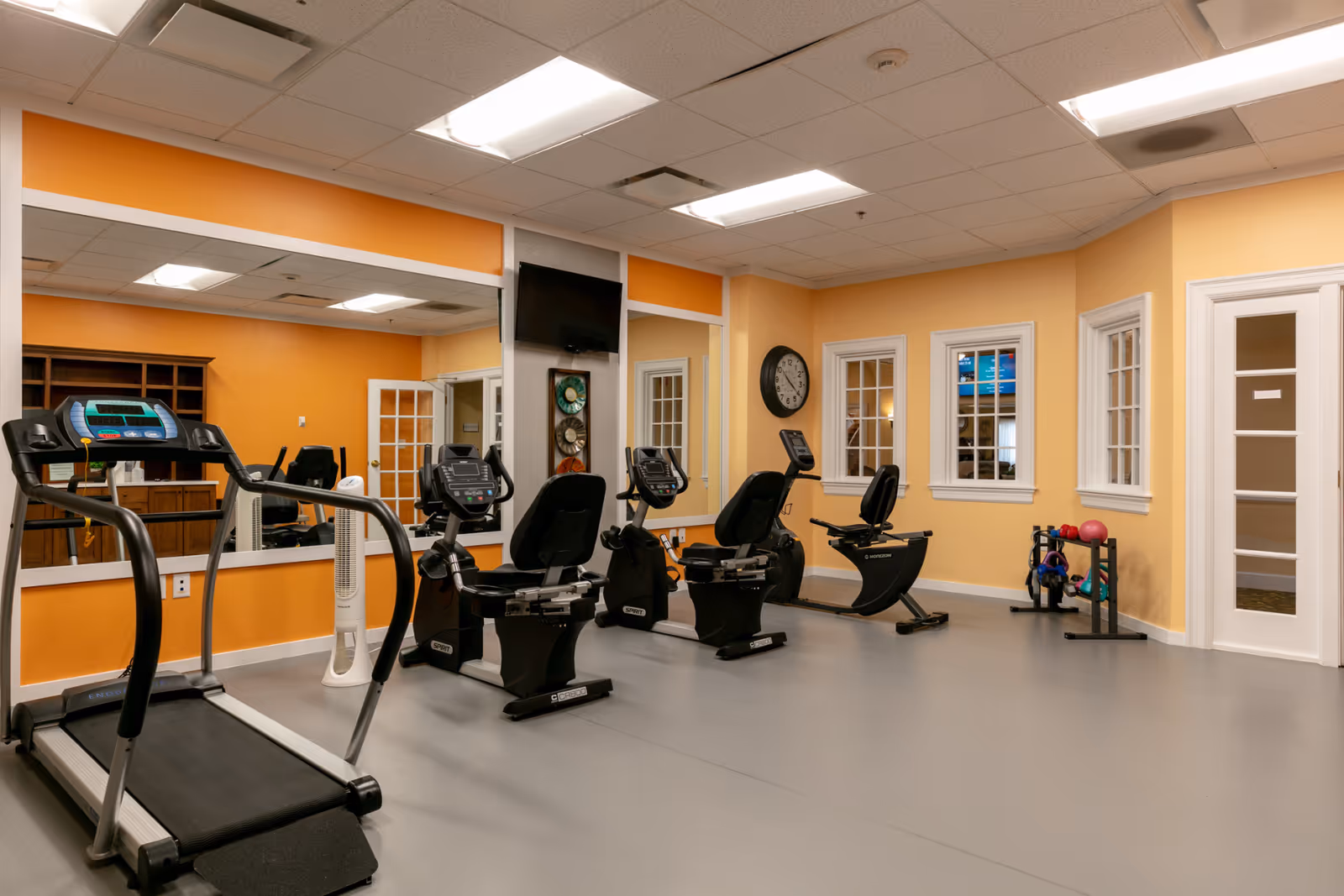 Interior fitness room with a treadmill, recumbent bikes, mirrors, and small free weights against orange walls.