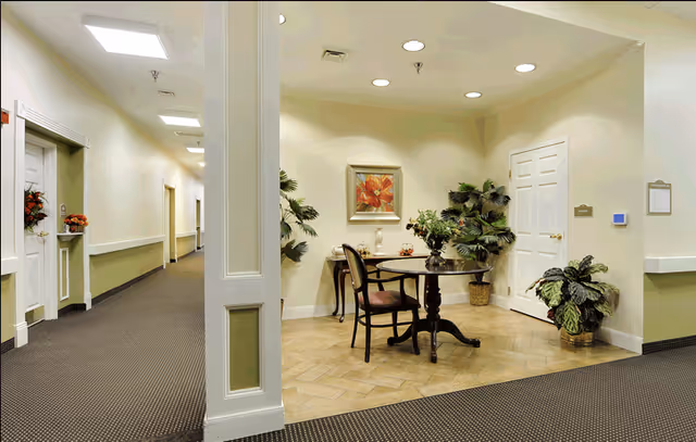 Interior view of a senior living facility hallway with beige walls and carpeted floors. To the right, there is a small seating area with a round wooden table, two chairs, potted plants, and a framed floral painting on the wall. The hallway extends to the left with several doors and handrails along the walls.