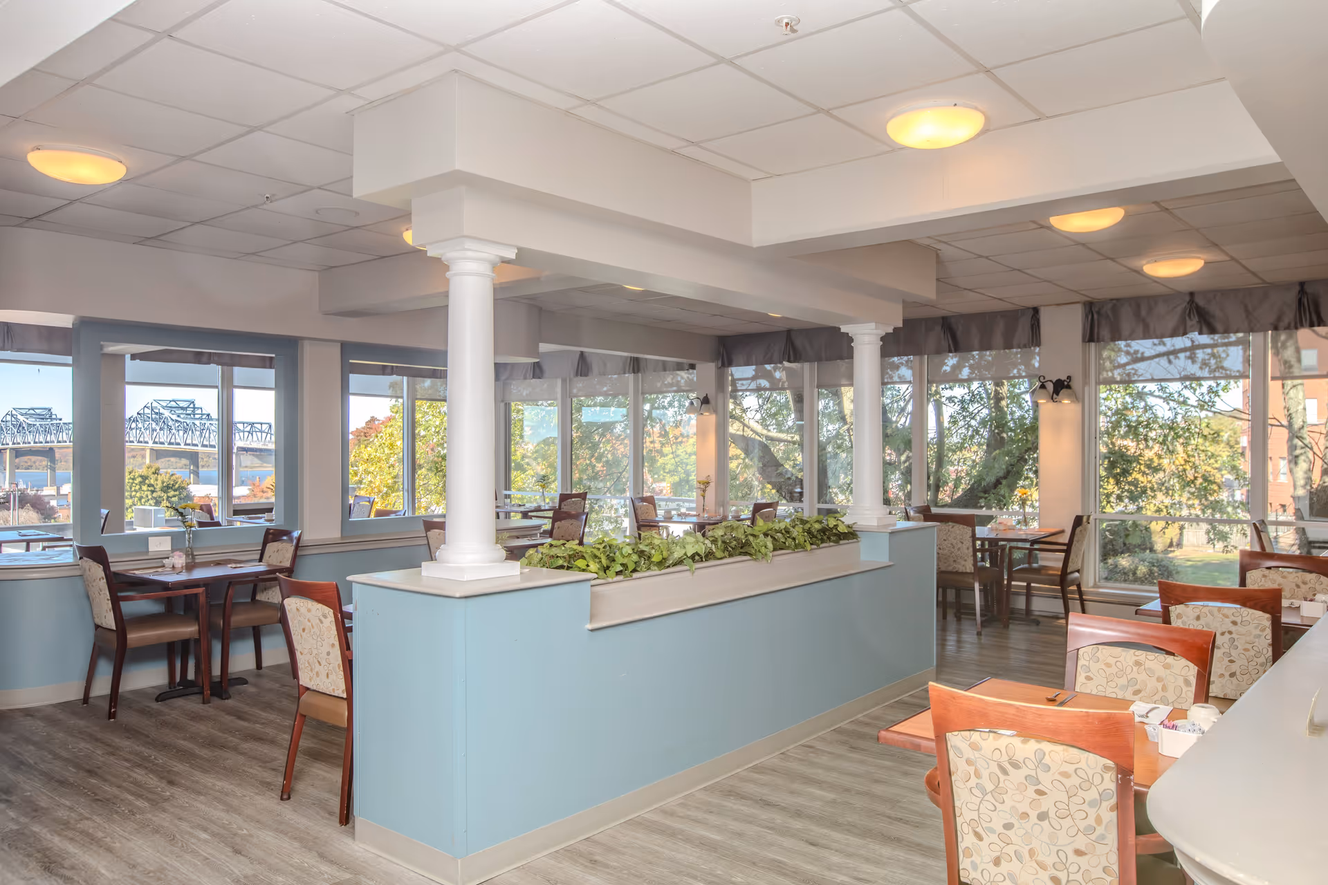 Bright dining area with multiple tables and chairs, large windows offering views of trees and a bridge outside, light blue half walls with white columns, and ceiling lights illuminating the space.