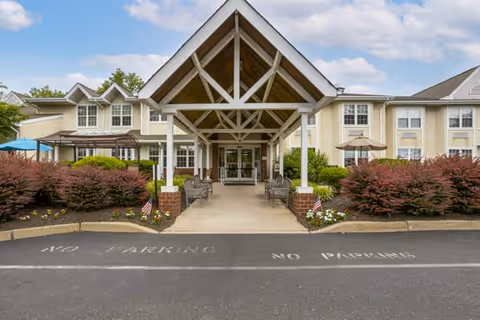 Front exterior view of Brookdale Northampton facility showing a covered entrance with a peaked roof supported by white beams. There are benches along the walkway leading to the glass doors. The building is two stories with multiple windows, surrounded by landscaped bushes and flowers. The sky is partly cloudy.
