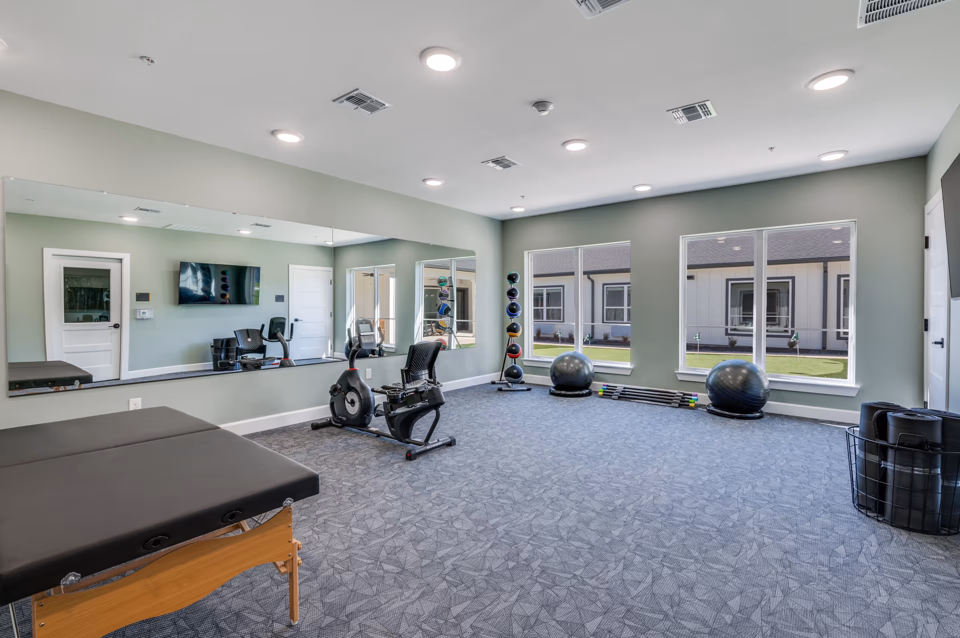 A bright exercise room with large windows showing an outdoor view. The room has a recumbent exercise bike, a massage table, a rack of colorful medicine balls, two large exercise balls, and several exercise bars. The walls are painted light green, and there is a large mirror on one side of the room reflecting the equipment and windows.