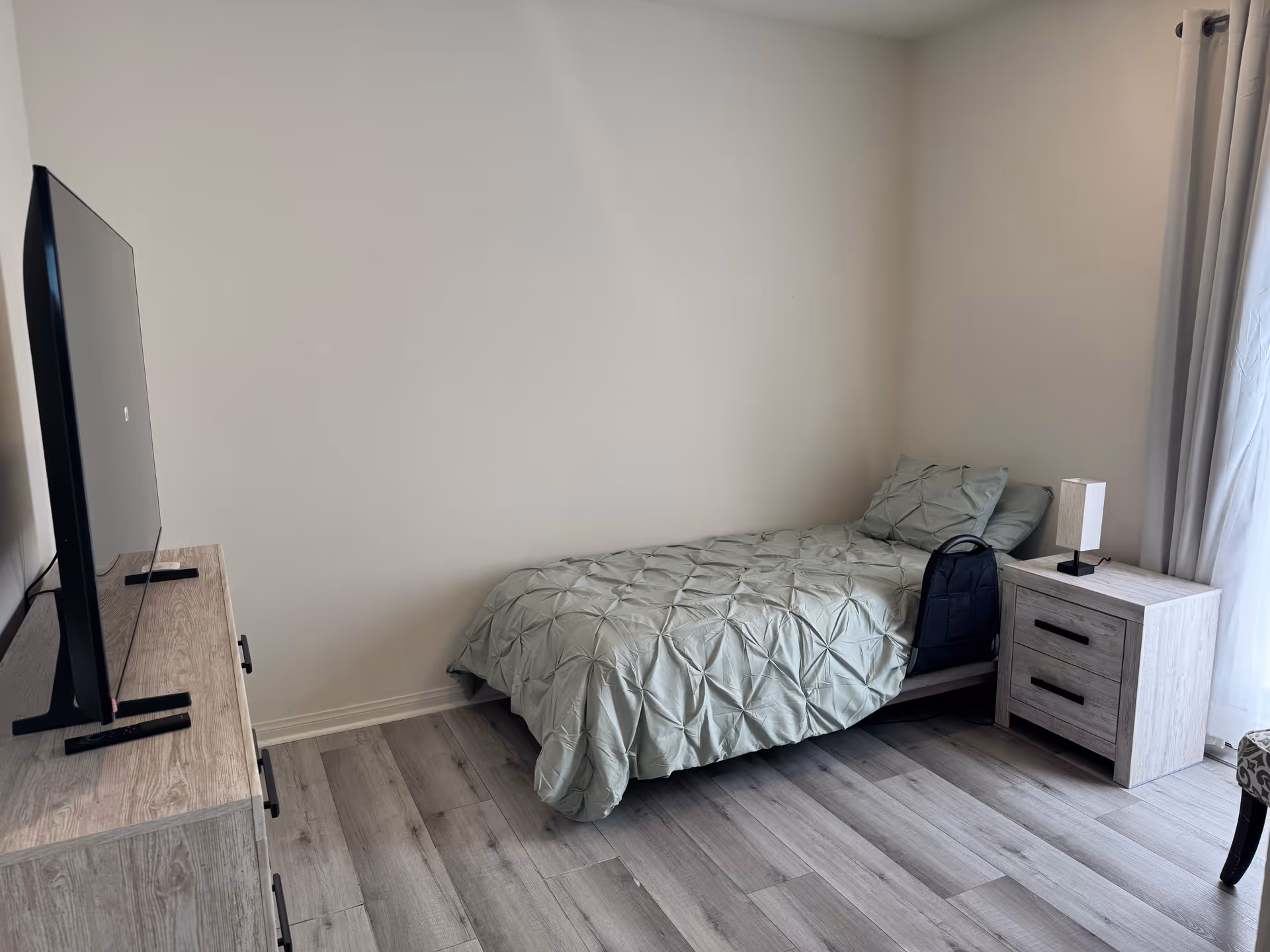 A small bedroom with a single bed covered in a light green textured comforter and matching pillow. Next to the bed is a light wood nightstand with two drawers and a small lamp on top. A black backpack is placed on the bed near the nightstand. Across from the bed is a light wood dresser with a flat-screen TV on top. The room has light-colored wood flooring and white walls, with a window covered by light curtains on the right side.