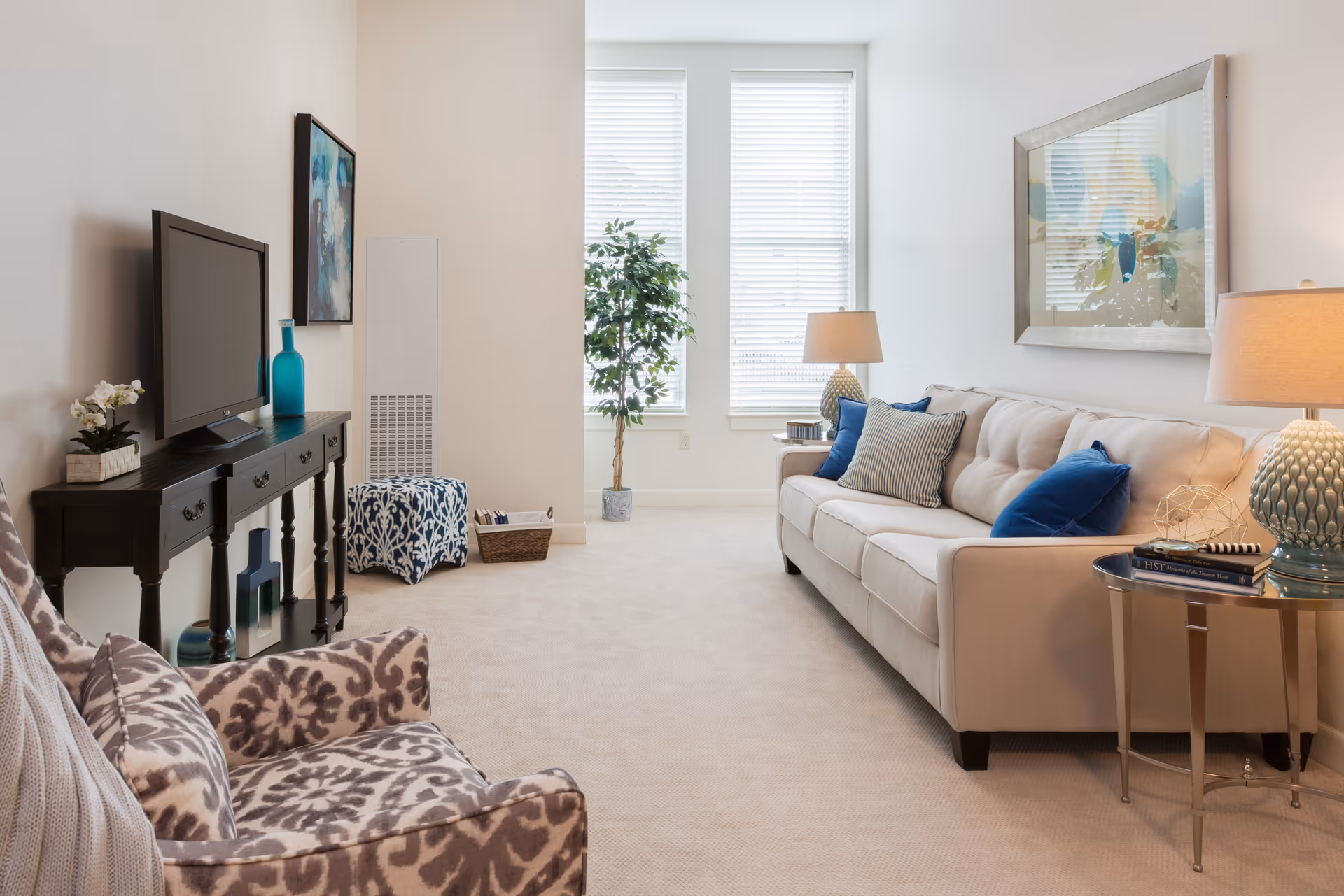 A bright and cozy living room with a beige sofa adorned with blue and striped pillows, a patterned armchair, a black console table with a flat-screen TV and decorative items, two large windows with white blinds, a potted plant, and two table lamps on side tables.