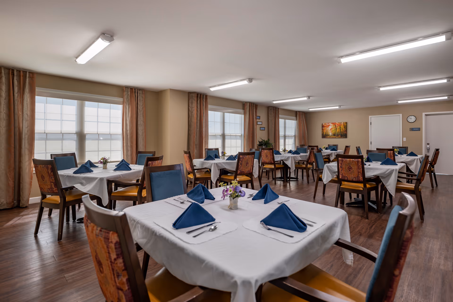 Communal dining room with multiple tables set with white tablecloths, folded blue napkins, and wooden chairs.