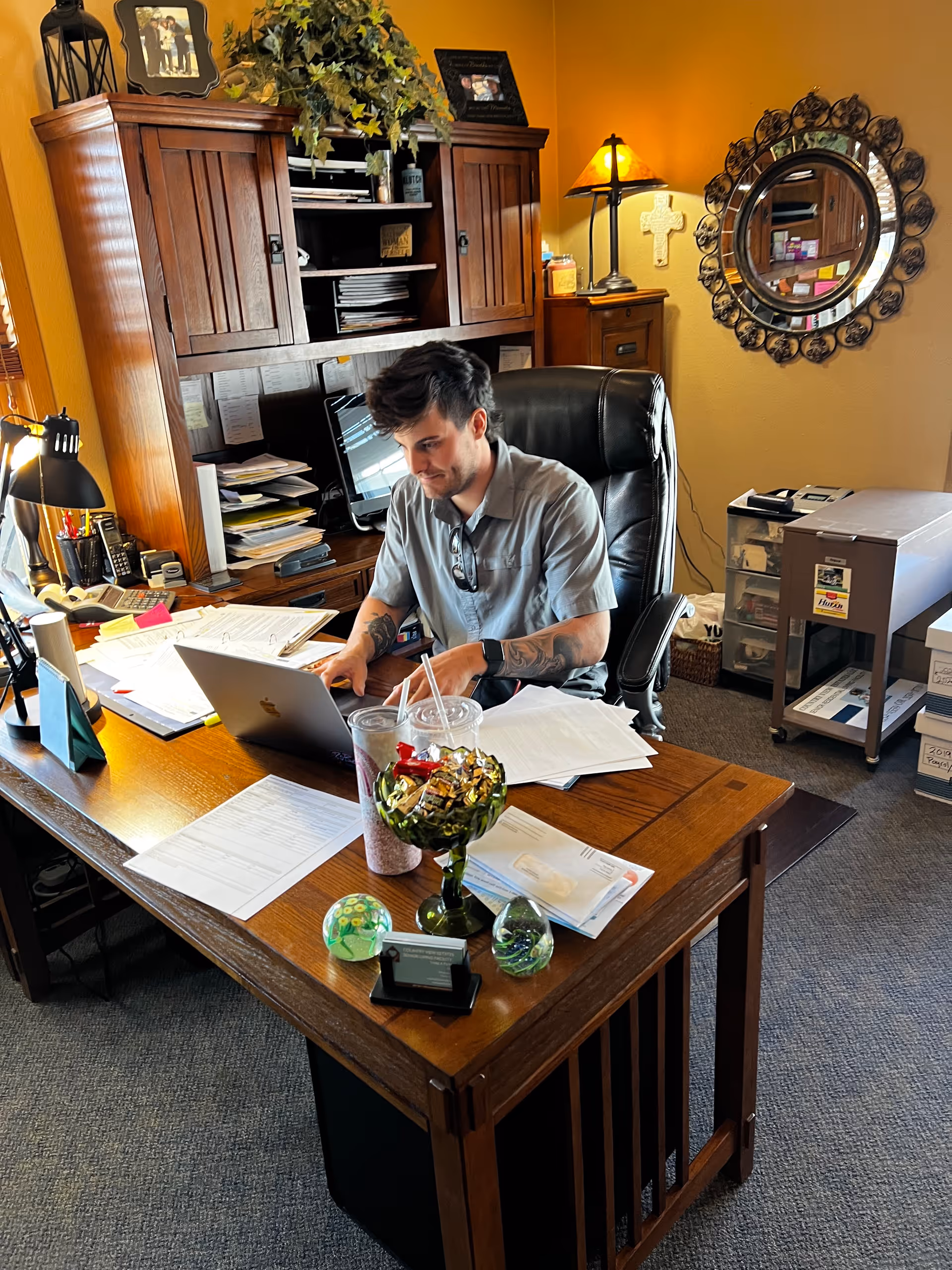 A man with tattoos on his arms is sitting at a wooden desk in an office, working on a laptop. The desk is cluttered with papers, two cups with straws, a decorative glass bowl filled with wrapped candies, and other small items. Behind him is a wooden hutch with shelves holding files and decorative plants. The room has warm yellow walls, a round decorative mirror, a table lamp, and filing cabinets.