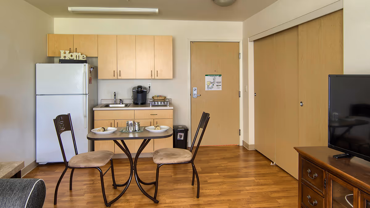 A small kitchen and dining area in a senior living facility apartment. The kitchen has light wood cabinets, a white refrigerator, a sink, and a coffee maker on the counter. In front of the kitchen is a small dining table set for two with plates and napkins, accompanied by two chairs. To the right, there is a wooden TV stand with a flat-screen television. The floor is wood, and there is a door with an evacuation plan posted on it. Sliding closet doors are visible next to the door.