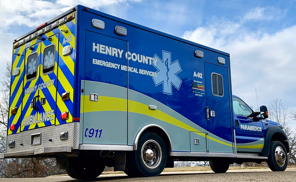 A Henry County blue and yellow ambulance parked outdoors under a partly cloudy sky.
