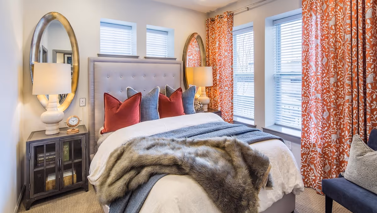 A cozy bedroom with a tufted headboard bed adorned with red and gray pillows, a gray blanket, and a fur throw. There are two oval mirrors on the walls behind matching bedside tables with lamps. The room has three windows with white blinds and orange floral curtains, and a blue chair with a gray pillow is partially visible.