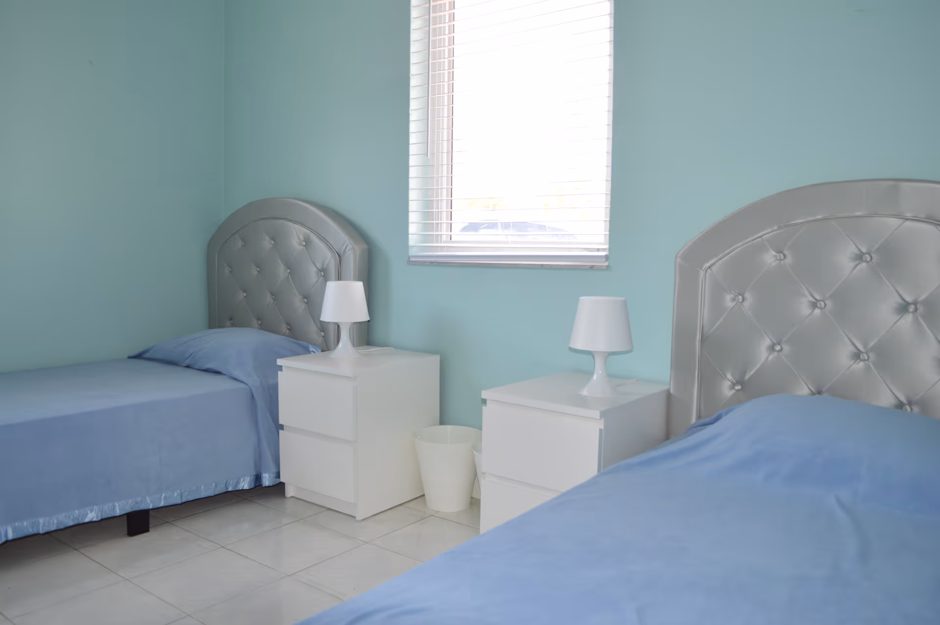 A bedroom with two single beds featuring tufted silver headboards and blue bedspreads. Between the beds are two white nightstands, each with a white lamp. A window with blinds is centered on the light blue wall behind the nightstands.