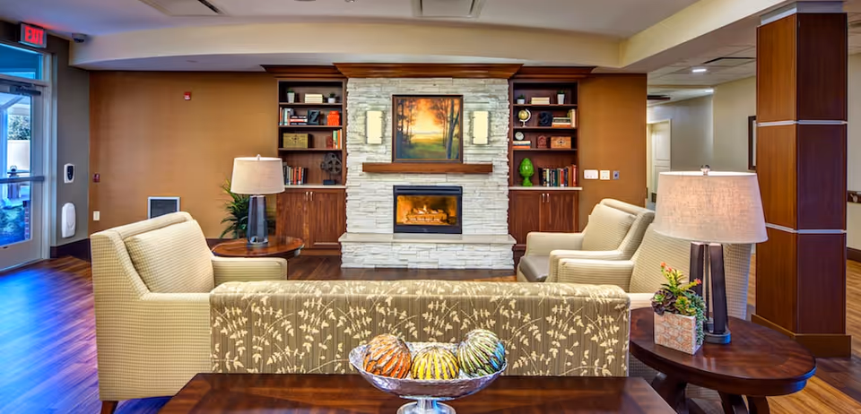 A cozy living room area in a senior living facility featuring a stone fireplace with a framed painting above it, flanked by wooden bookshelves filled with books and decorative items. The room has comfortable beige armchairs and a patterned sofa facing the fireplace. There are two side tables with lamps and a decorative bowl with colorful spheres on a wooden table in the foreground. The floor is wooden, and there is an exit door visible on the left side.