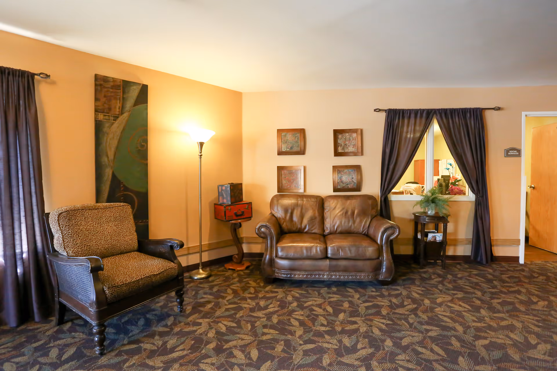 A cozy living room area with a brown leather loveseat, a patterned armchair, a floor lamp, and a small side table with a plant. The walls are painted beige and decorated with four square framed artworks. There are dark curtains on the windows and a door leading to another room.