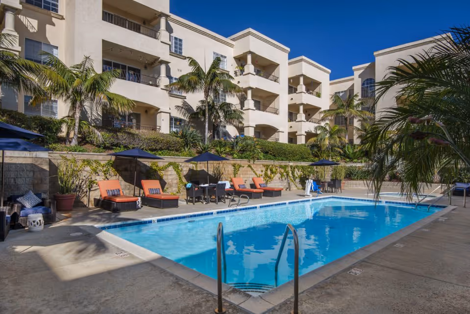 Outdoor swimming pool area at La Vida Del Mar facility with lounge chairs, umbrellas, palm trees, and a multi-story beige building in the background under a clear blue sky.