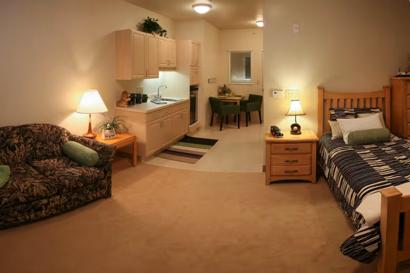 Studio-style senior living interior with a bed, nightstand, sofa, kitchenette, and small dining table.
