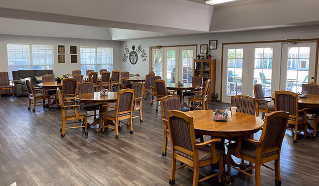 A spacious dining room with multiple round wooden tables and matching chairs arranged neatly on a wood floor. Large windows and glass doors allow natural light to fill the room. There is a wall clock and decorative items on the walls, and a small bookshelf near the doors.