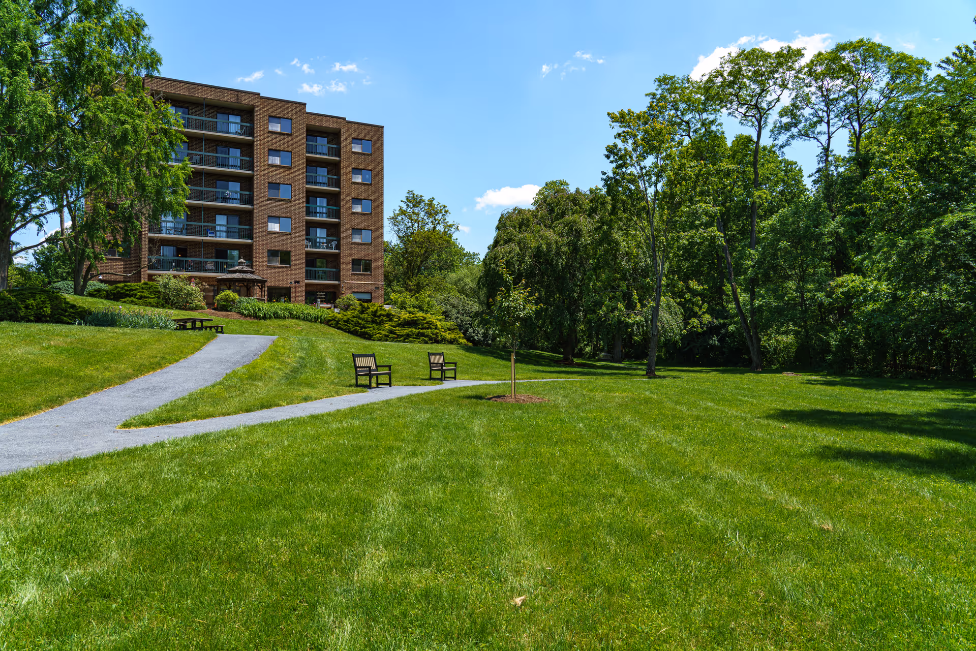A large green lawn with a paved walking path leading to a multi-story brick building with balconies. There are two benches along the path and several trees surrounding the area under a clear blue sky.