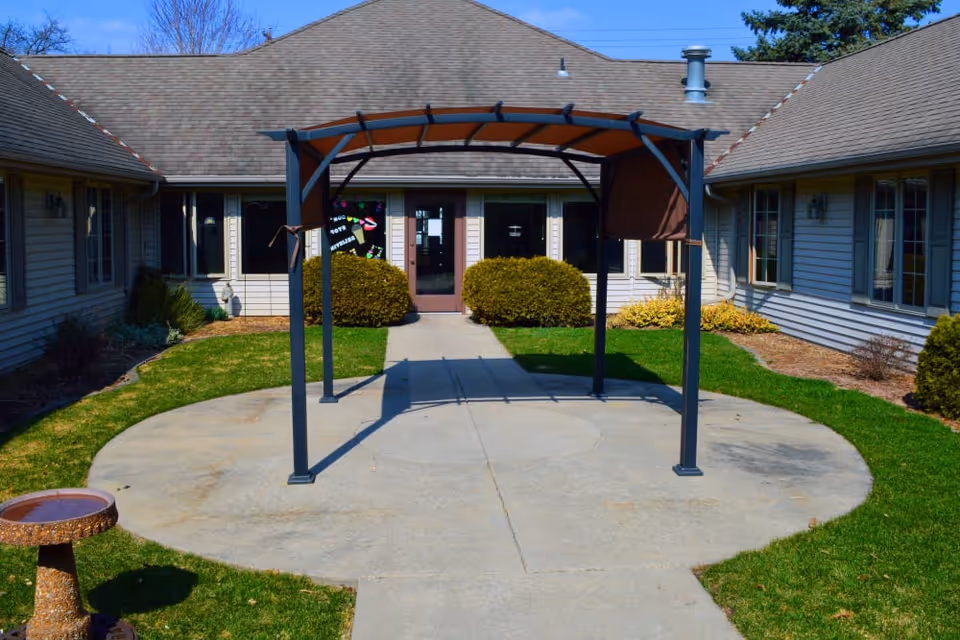 Small courtyard with a metal pergola over a circular concrete pad leading to the entrance of a single-story memory care building flanked by shrubs.