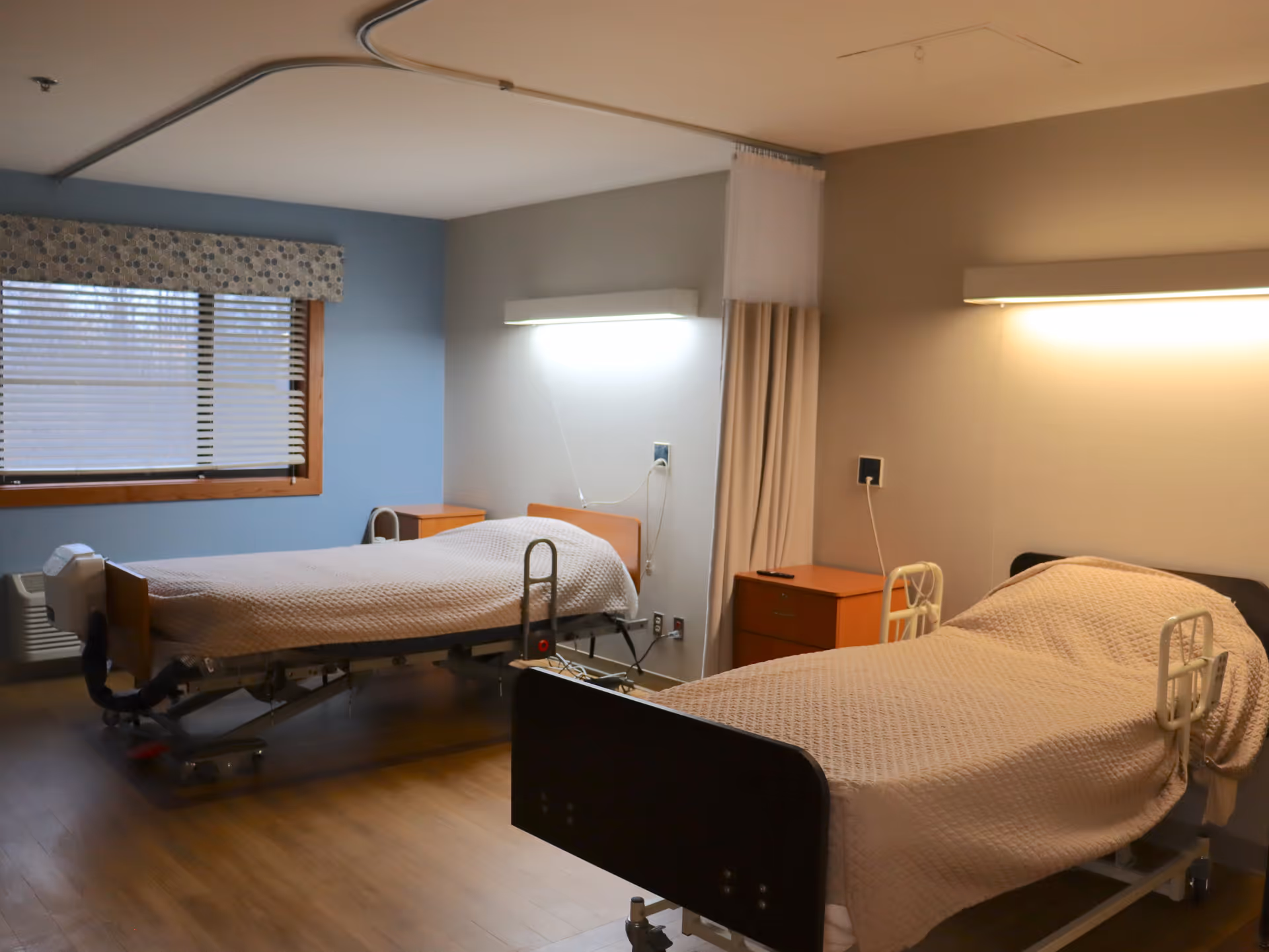 A room with two hospital-style beds covered with beige blankets, each with side rails. The room has wooden flooring, a window with blinds and a patterned valance, two wooden nightstands, and wall-mounted lights above each bed. A ceiling-mounted curtain track with a partially drawn privacy curtain is visible between the beds.