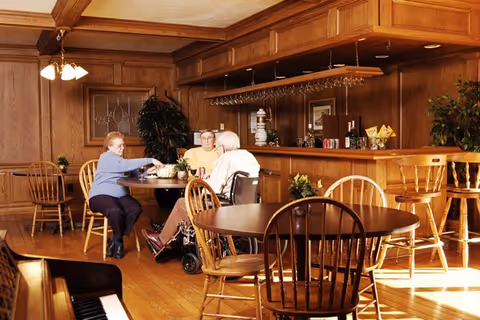Three elderly people sitting around a wooden table in a cozy room with wood-paneled walls and a bar counter in the background. One person is in a wheelchair, and the others are seated on wooden chairs. The room is warmly lit with a chandelier and has several plants and framed pictures on the walls.