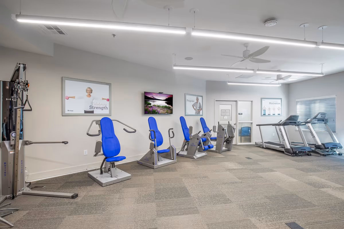 A fitness room with exercise equipment including seated strength machines with blue seats and two treadmills near a window. The walls have motivational posters and a TV mounted in the center. The room is well-lit with ceiling lights and fans.