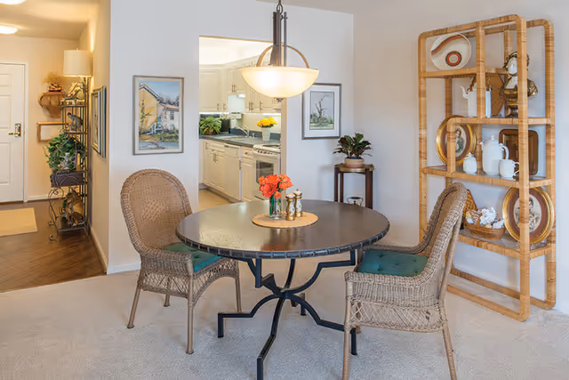 A cozy dining area with a round dark wooden table and two wicker chairs with green cushions. The table has a small centerpiece with orange flowers and salt and pepper shakers. Behind the table is a wooden shelving unit displaying decorative plates and white ceramic items. The dining area opens into a kitchen with white cabinets and appliances. The walls are decorated with framed artwork, and there is a hallway leading to the entrance door with a floor lamp and plants on a metal shelf.