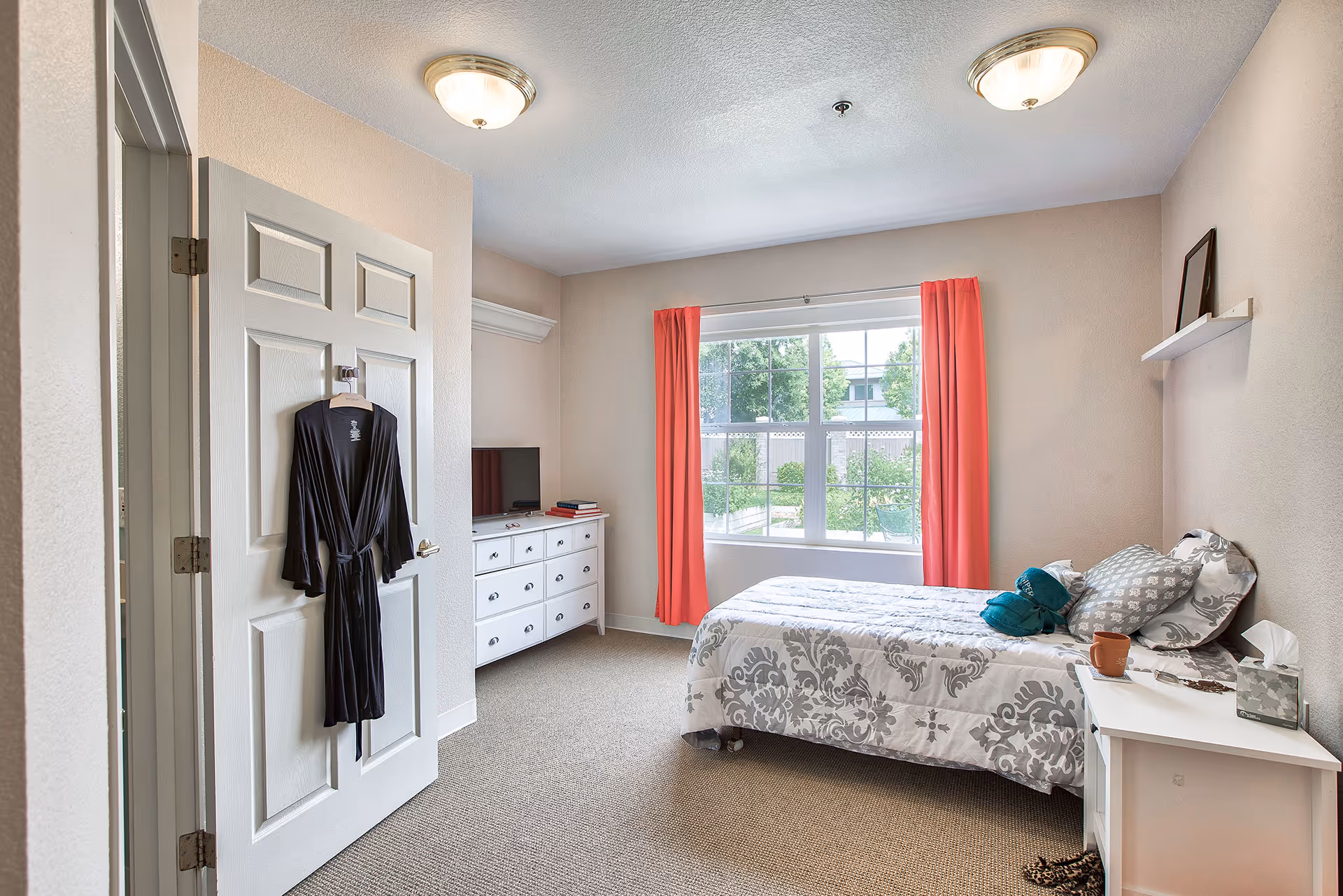 A bright bedroom with a single bed covered in a white and gray patterned comforter, a white nightstand with a tissue box and a cup, a white dresser with a TV and books, coral curtains framing a large window showing a garden outside, and a black robe hanging on the door.
