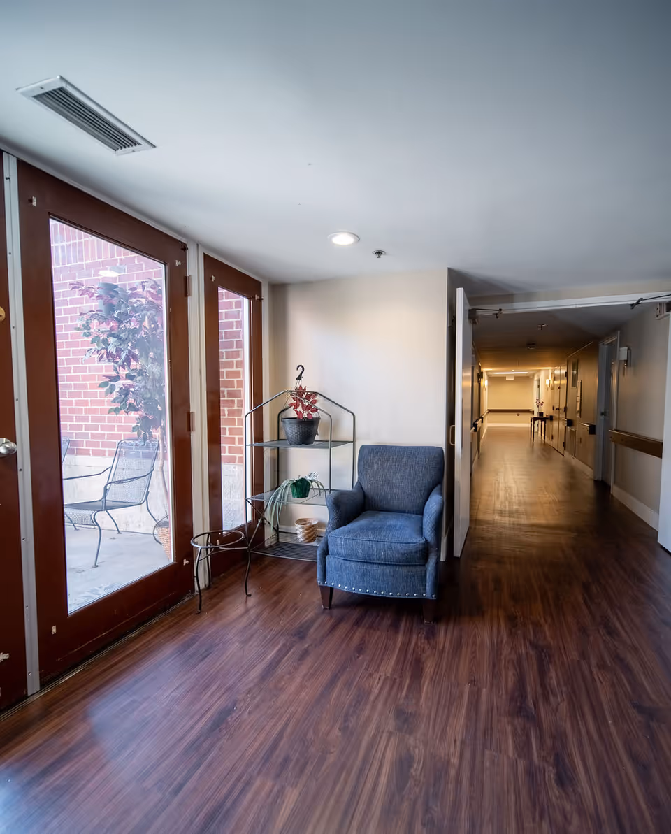 A hallway with wooden flooring featuring a blue upholstered armchair and a metal shelf with potted plants. To the left, there are glass doors leading to an outdoor patio with metal chairs and a brick wall. The hallway extends into the distance with doors and handrails along the walls.