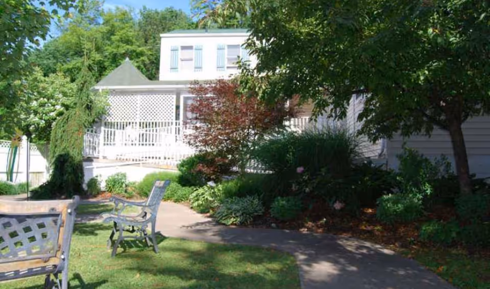 Landscaped outdoor seating area with metal benches and chairs in front of a white residence with a covered porch.
