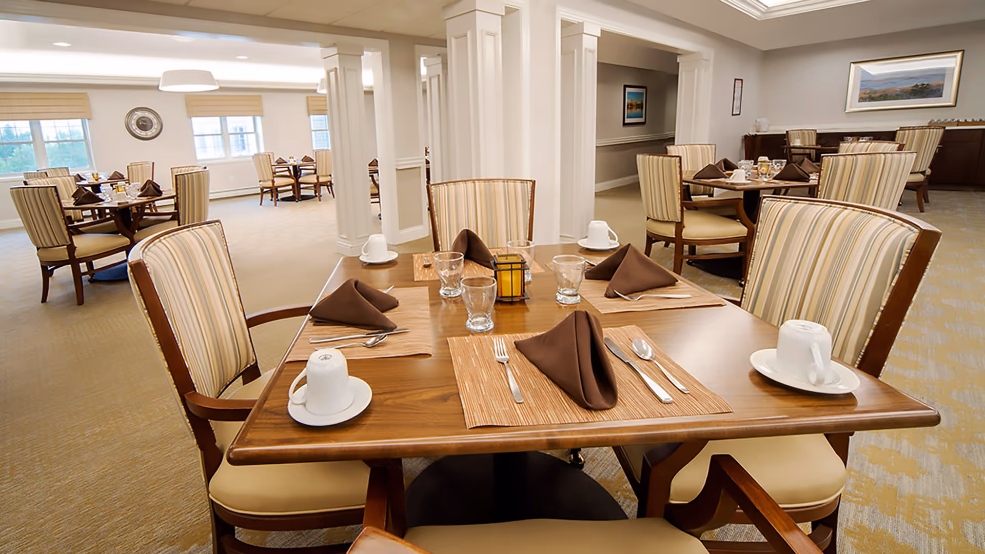 Well-lit dining room with wooden tables set with napkins, cups, glasses, and striped upholstered chairs.