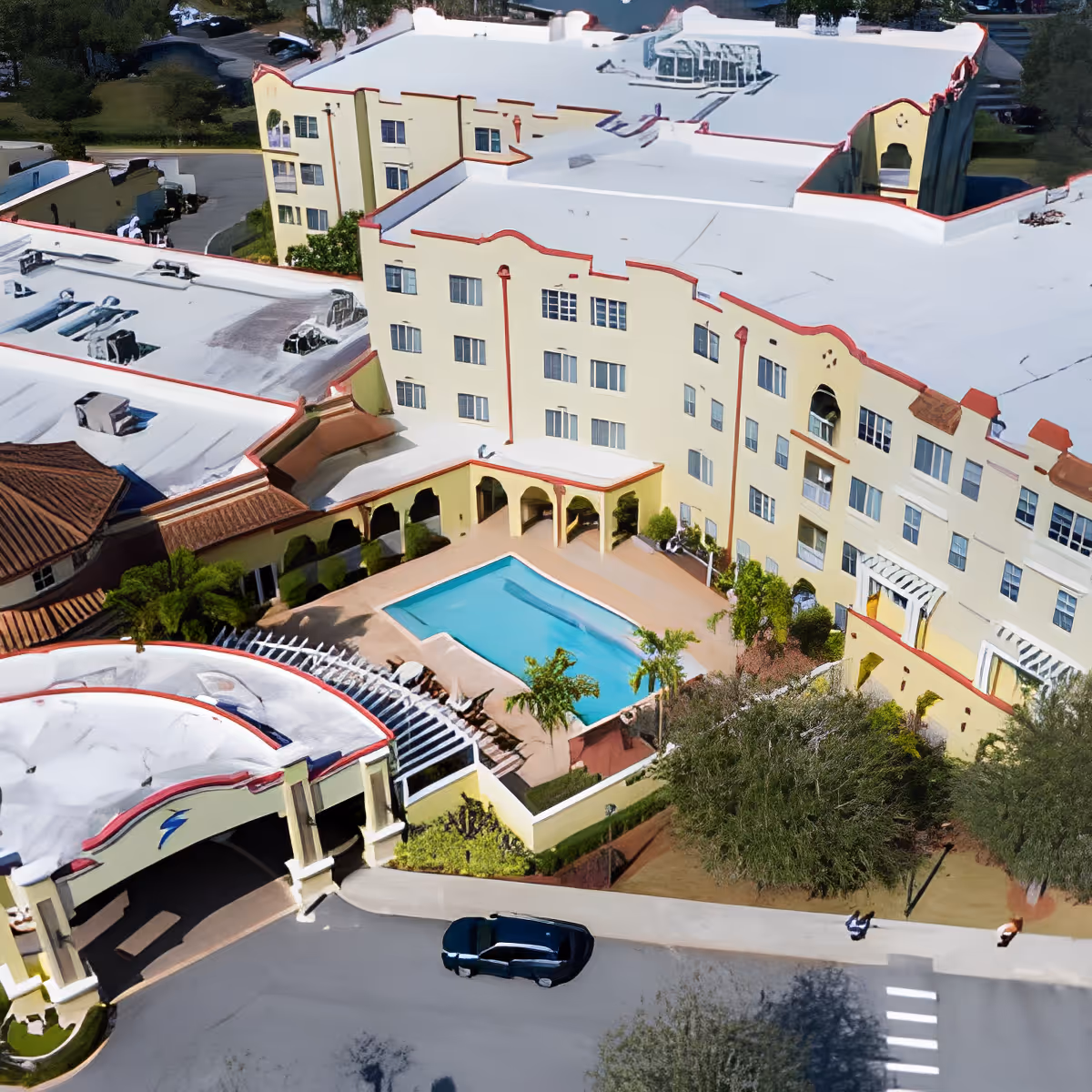 Aerial view of Discovery Village At Melbourne showing a multi-story yellow building with red trim surrounding a central outdoor swimming pool. The building has multiple windows and a covered entrance area. There are trees and greenery around the building and a few cars parked nearby.