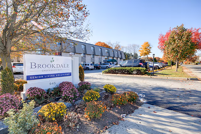 Exterior view of a senior living facility named Brookdale Greenwich Bay with a parking lot, autumn trees, and colorful flowers in the foreground.