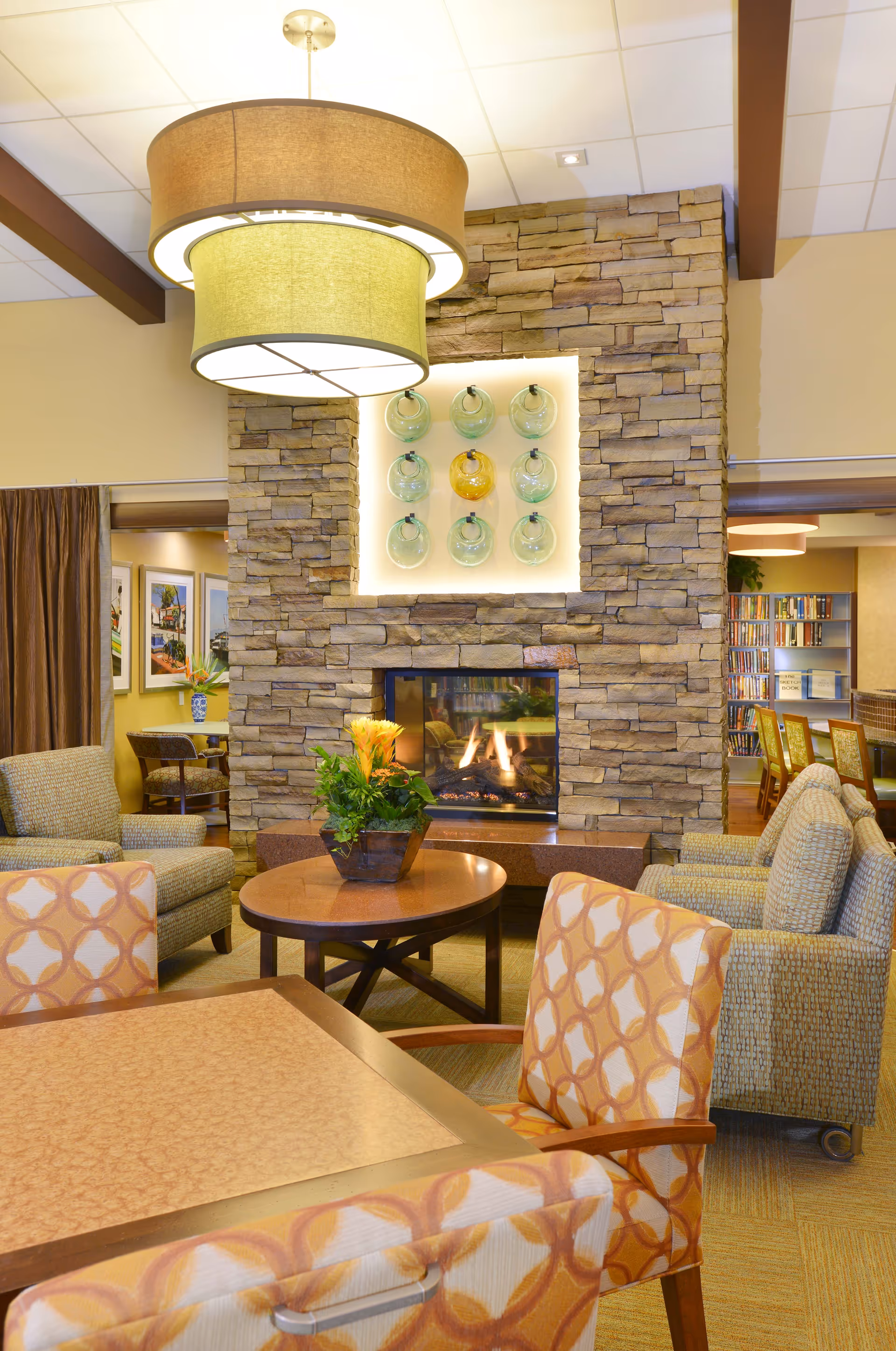 A cozy living room area in a senior living facility featuring a stone fireplace with a decorative wall niche above it containing glass art pieces. The room has comfortable upholstered chairs with patterned fabric around a wooden table, a round coffee table with a plant centerpiece, and warm lighting from a large ceiling fixture. In the background, there is a glimpse of a bookshelf and additional seating areas.