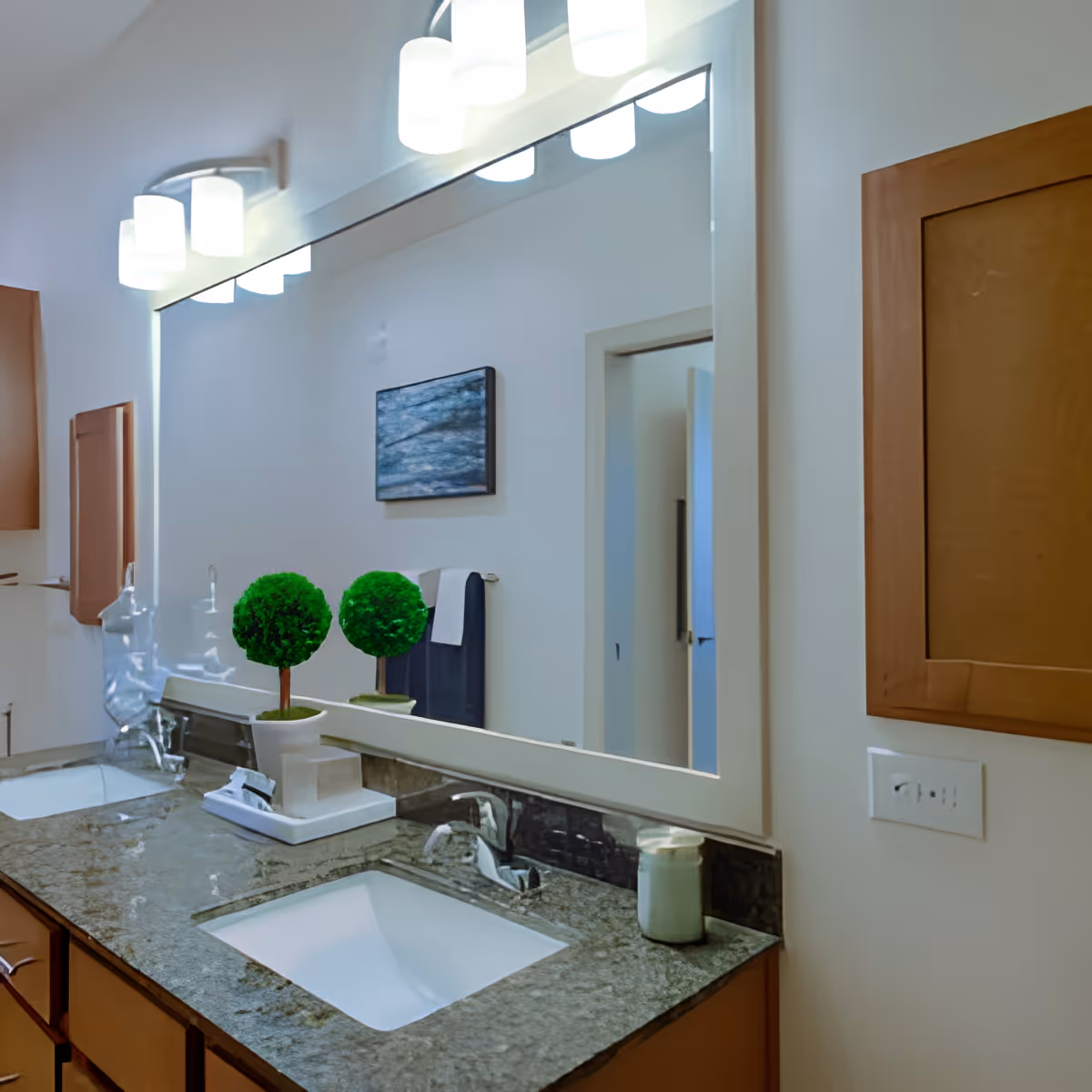 A modern bathroom vanity with a granite countertop featuring two sinks, a large mirror above, two small decorative potted plants, a soap dispenser, and a towel hanging on the wall. The room is well-lit with three light fixtures above the mirror and has wooden cabinets below the countertop.
