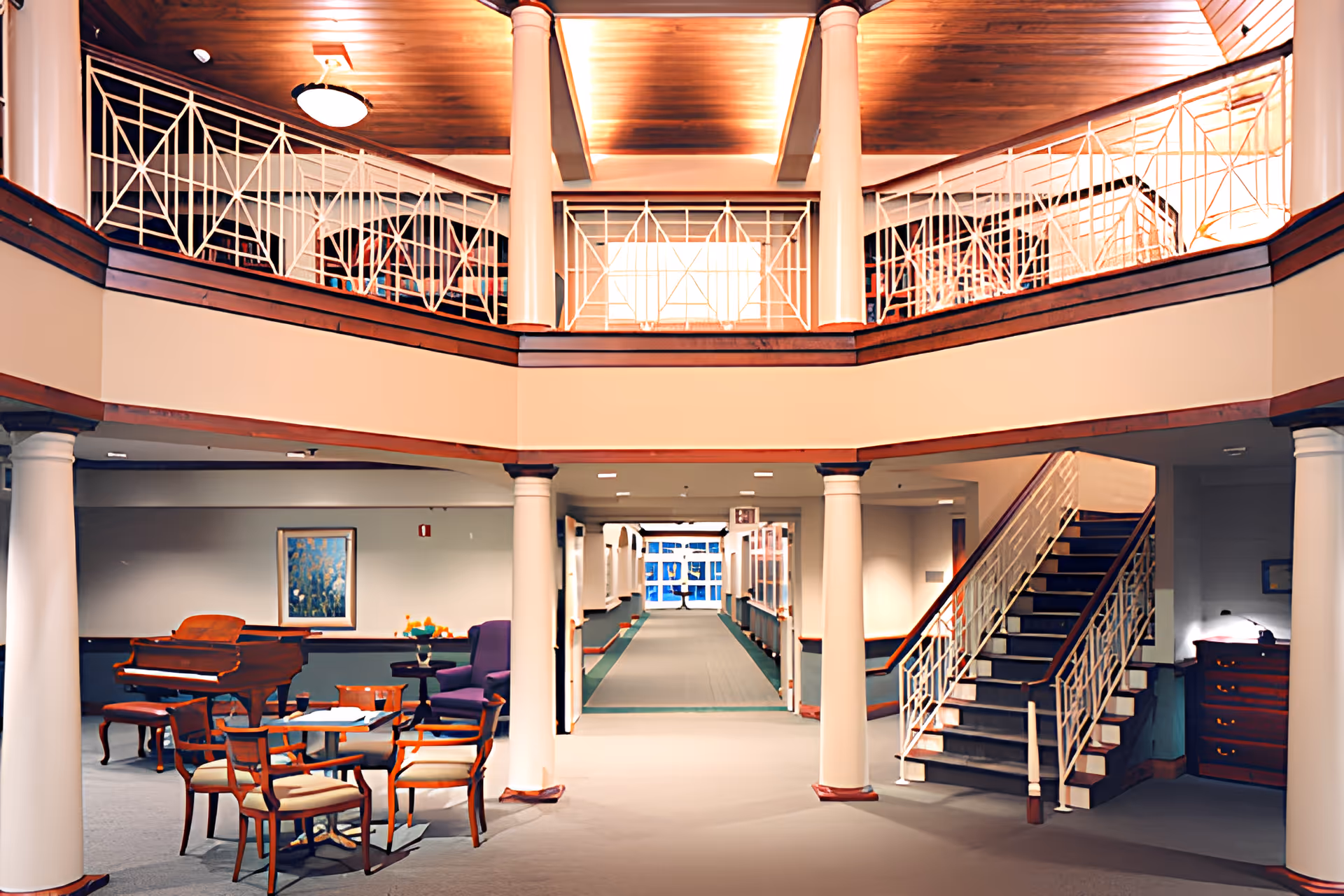 Interior view of a senior living facility lobby area with a staircase on the right, white columns, a seating area with chairs and a table, a grand piano, and a balcony with decorative railings above.