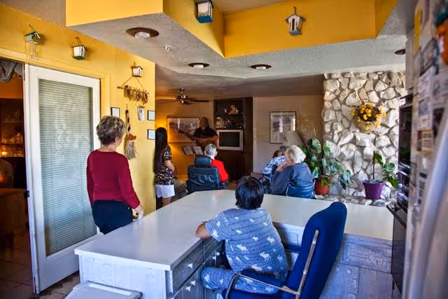 Several residents sit and stand in a bright communal living room with a large table and stone fireplace while someone plays guitar near a TV.