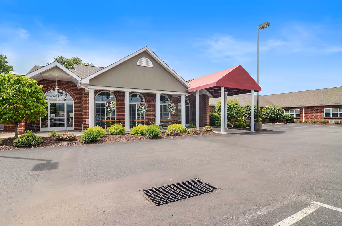 Front entrance of a brick senior living facility with a red covered drive-up canopy, large arched windows, and landscaped shrubs.