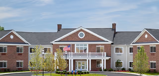 Front exterior view of a two-story senior living facility building named The Landing Of Brighton, featuring red brick and beige siding, white trim, a balcony with white railings, an American flag, and landscaped greenery with small trees and bushes.