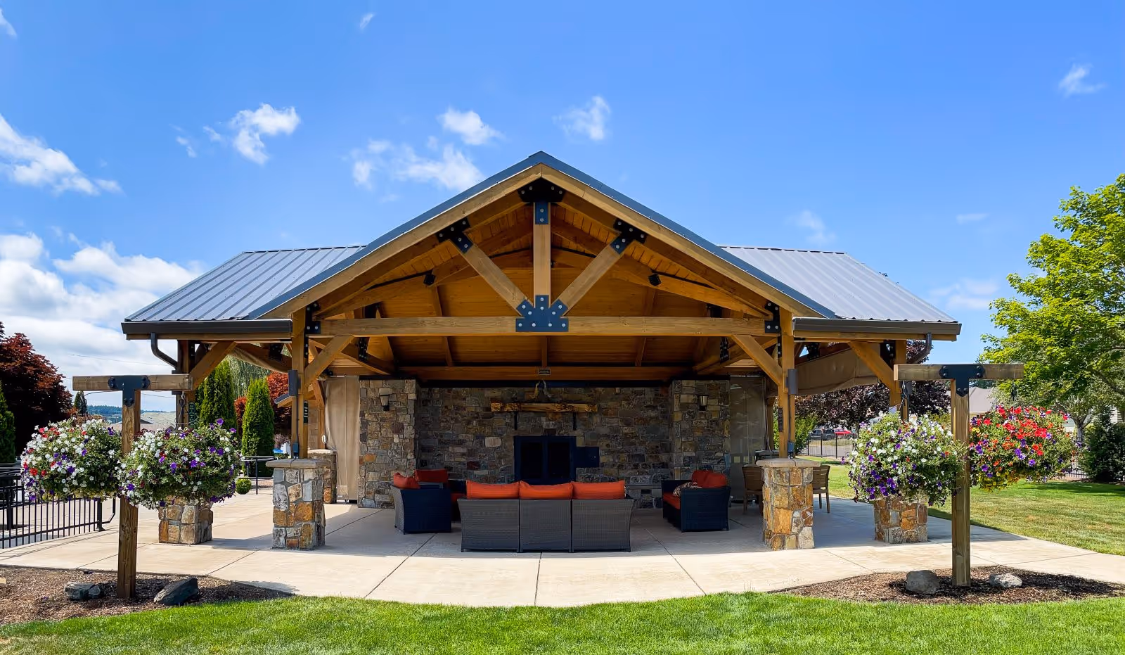 Outdoor covered seating area with a wooden roof structure and stone pillars, furnished with wicker sofas and orange cushions, surrounded by hanging flower baskets and green lawn under a blue sky with scattered clouds.
