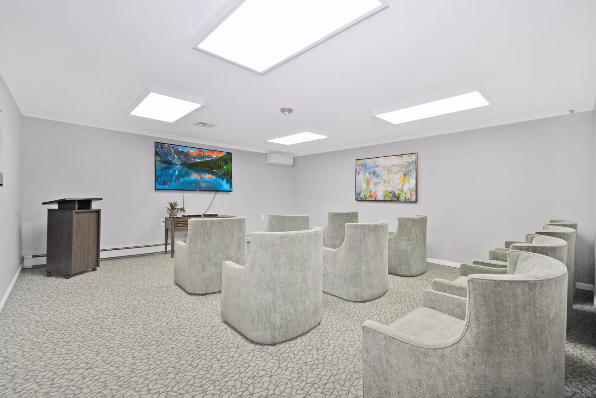 A small conference or presentation room with nine light gray upholstered chairs arranged in rows facing a wall-mounted flat screen TV displaying a scenic mountain and lake image. The room has light gray walls, a patterned carpet, a wooden podium on the left side, a small table under the TV with a plant, and a colorful abstract painting on the right wall. The ceiling has four large rectangular fluorescent lights.