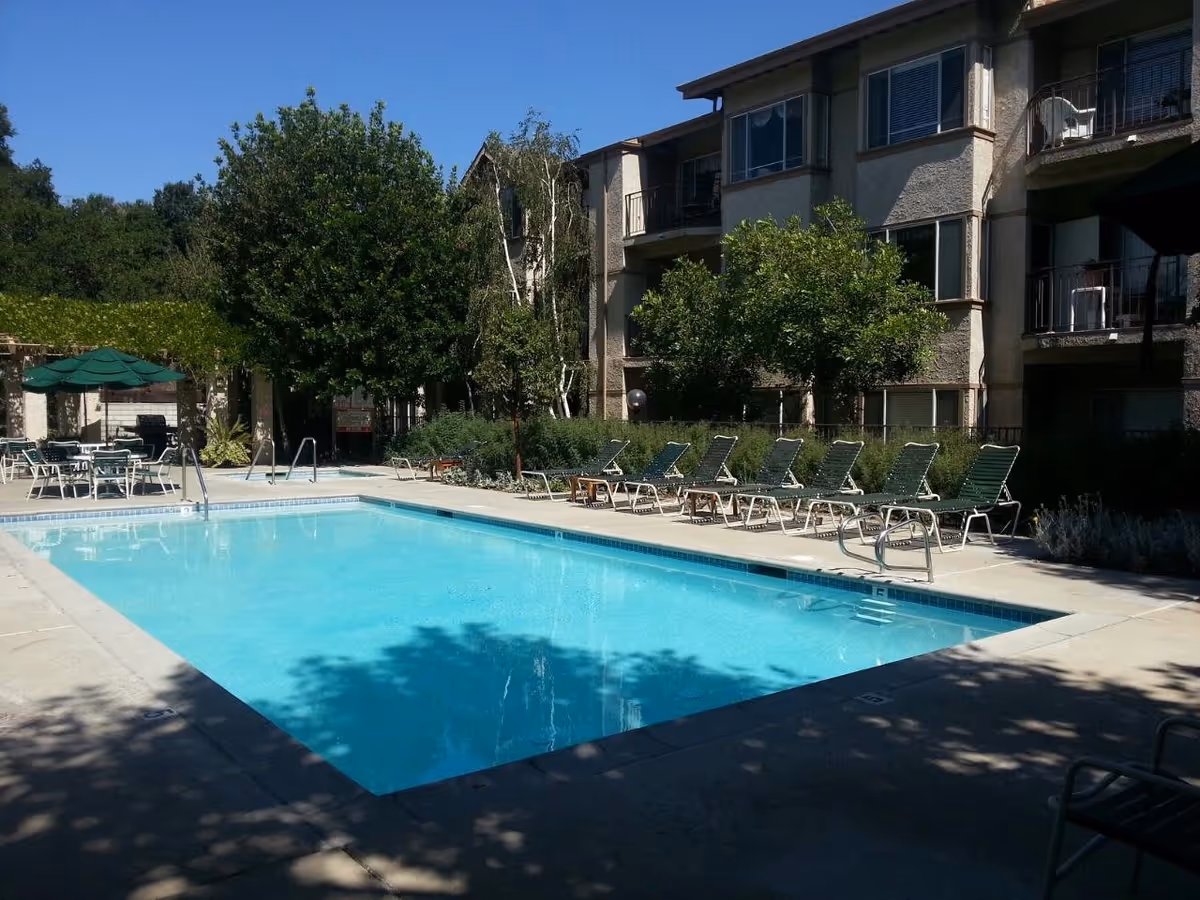 Outdoor swimming pool with clear blue water surrounded by lounge chairs and tables with green umbrellas. Trees and shrubs are around the pool area, and a multi-story residential building is visible in the background under a clear blue sky.