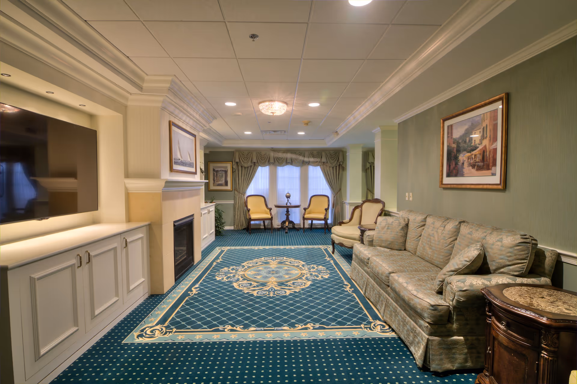 A cozy living room with a patterned blue carpet, a greenish sofa, an armchair, a wooden side table, and two yellow cushioned chairs near a window with green curtains. There is a fireplace with a painting above it and a large flat-screen TV mounted on the wall opposite the sofa.