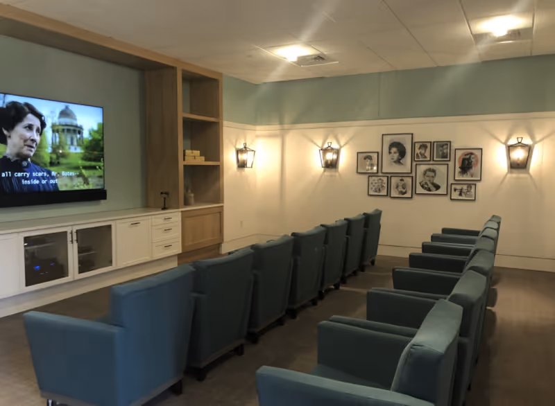 A small theater-style room with two rows of blue upholstered armchairs facing a wall-mounted flat screen TV. The TV is showing a scene with a woman speaking and subtitles visible. The room has light blue and white walls, wall-mounted lantern-style lights, and a gallery of black and white framed portraits on one wall. There is a built-in cabinet and shelving unit beneath the TV.