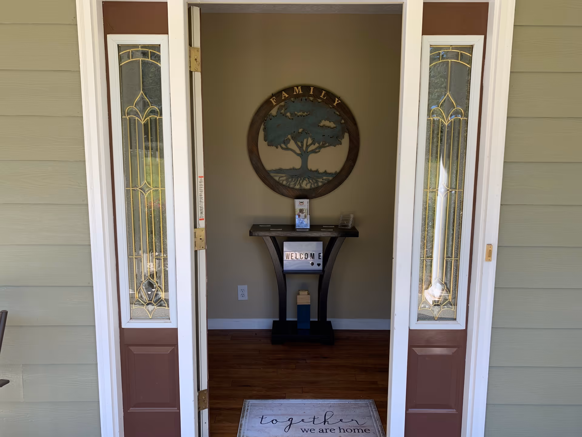 Open front door revealing an entry foyer with a welcome mat, a small console table and a round 'Family' wall sign.