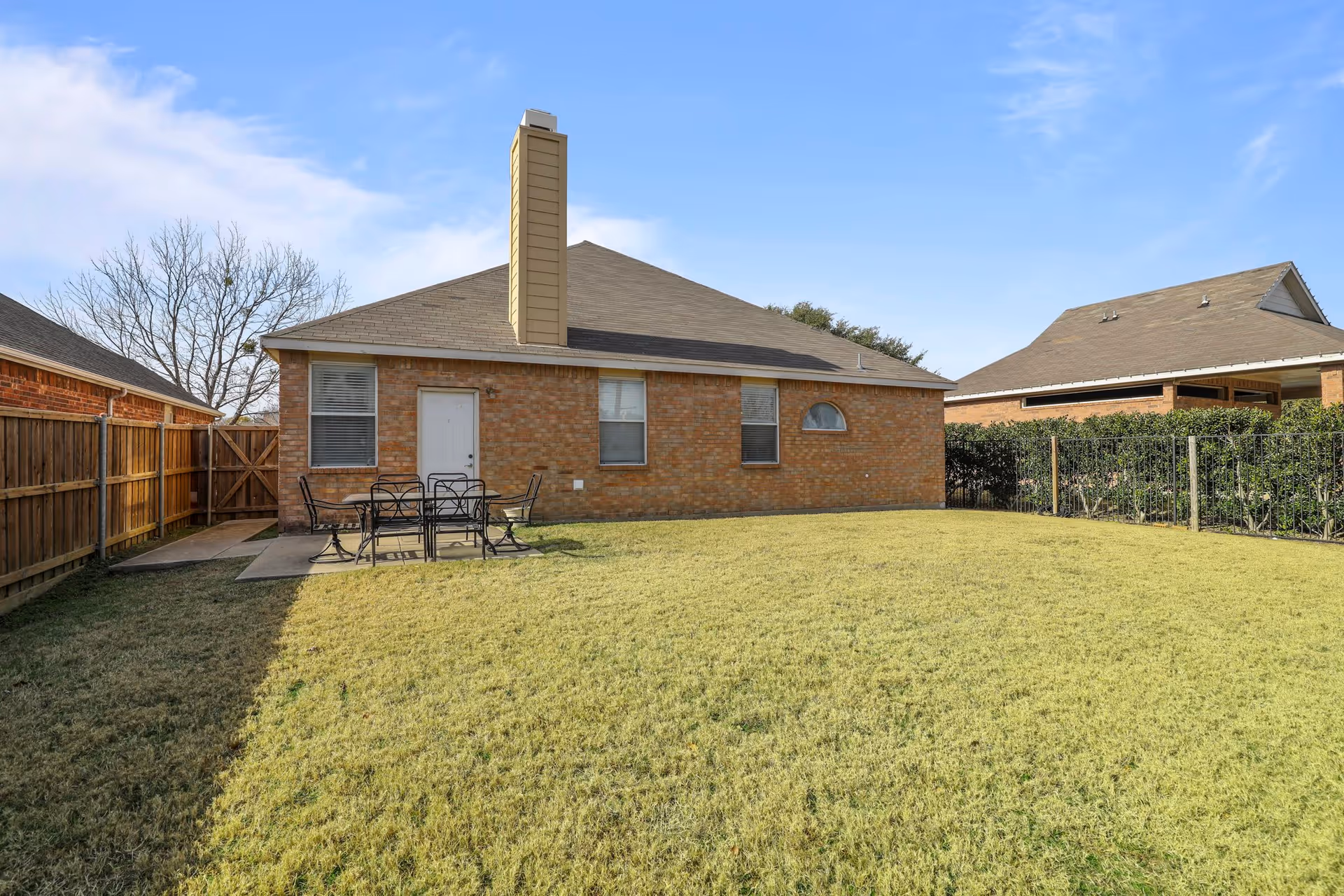 A backyard with a green lawn and a small concrete patio area with a black metal table and four chairs. The house is made of brick with three windows and a white door. There is a wooden fence on the left side and a hedge fence on the right side. The sky is clear with a few clouds.