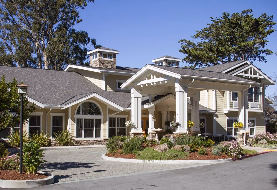 Exterior view of a senior living facility building with beige siding, white trim, and a covered entrance supported by large white columns. The building is surrounded by landscaped greenery, flowers, and trees under a clear blue sky.