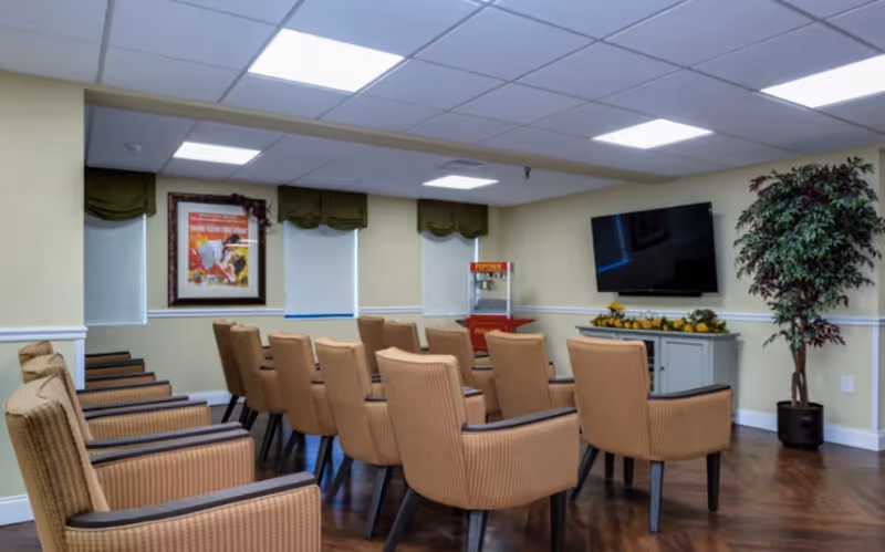 A small theater-style room with three rows of beige cushioned chairs facing a wall-mounted flat screen TV. The room has light yellow walls, three windows with green valances, a popcorn machine in the corner, a framed poster on the wall, and a large potted plant near the TV.