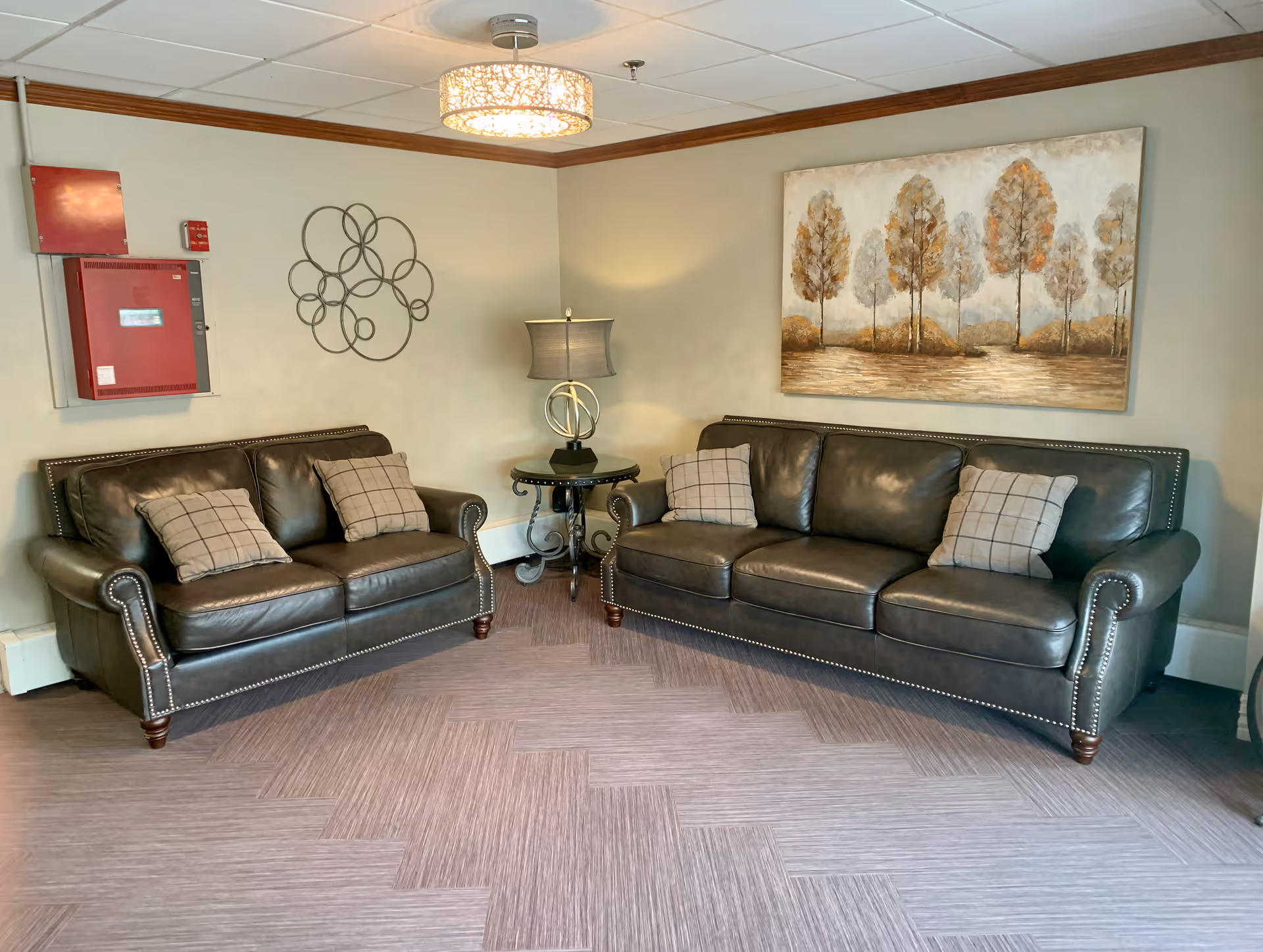 A cozy sitting area with two dark leather sofas and plaid pillows flanking a small round side table and lamp beneath a landscape painting.