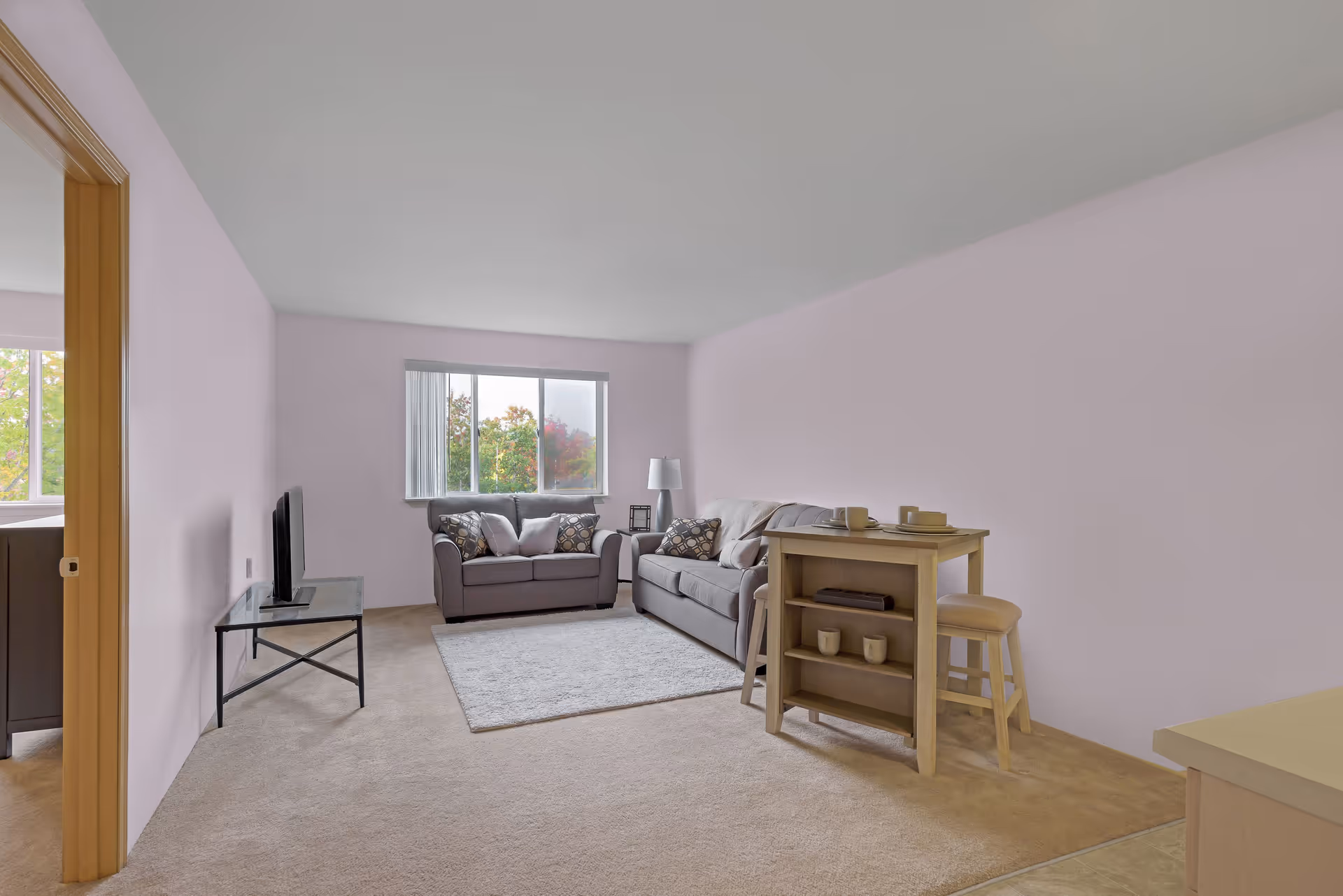 A living room with two gray sofas, a small wooden table with two stools, a floor lamp, a TV on a glass stand, and a window showing trees outside. The walls are painted light purple and the floor is carpeted.