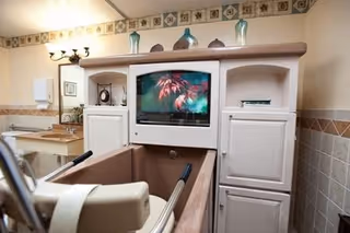 A bathroom in a senior care facility featuring a specialized bathtub with handrails and a TV screen mounted above it. The room has beige tiled walls with decorative border tiles near the ceiling, a sink with a mirror and light fixture, and cabinets for storage.